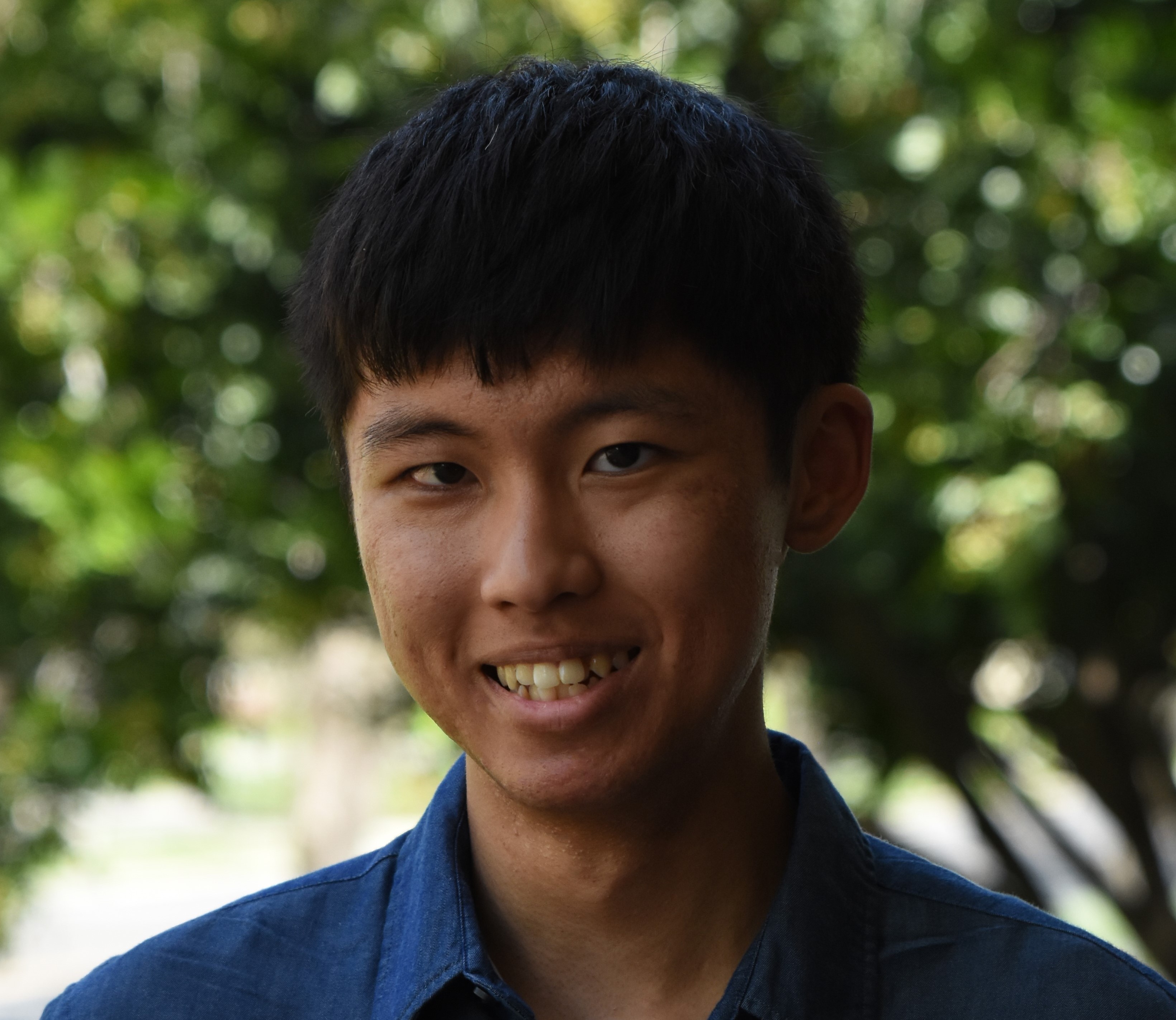 A photo of Gene, a Korean-American man smiling against a background with green trees. He has thick straight black hair that is longer on the front and shorter on the sides. He is wearing a blue collared shirt, and his dark brown eyes are smizing at the camera.